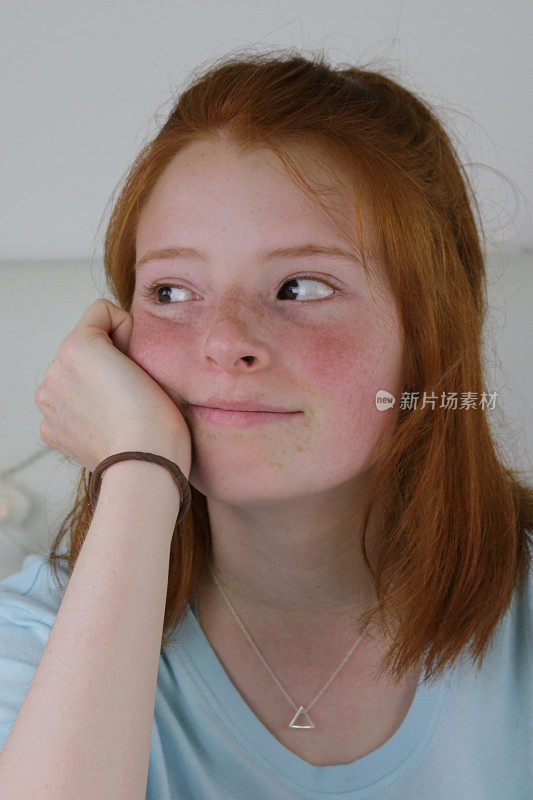 这是一个14 / 15岁的红发少女的照片，她皮肤苍白，脸上有雀斑，脸颊通红，坐在卧室里，看起来厌倦了，下巴靠在手上，看起来闷闷不乐